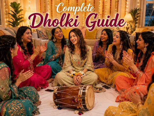 Women in bright shalwar kameez and sharara sets sitting cross-legged on a sheet-covered living room floor at a UK dholki, clapping around a dholak drum.